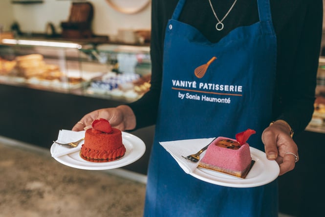A person in a blue Vaniyé Pâtisserie apron holding two small plates with one boutique French tart on each.