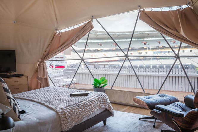 Glamping accommodation with bed and chair and window looking out across major sports stadium in Auckland