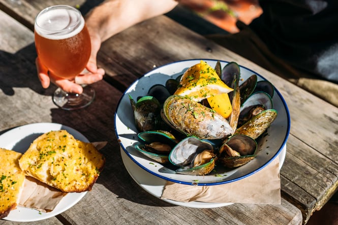 A man holding a glass of beer next to a bowl of mussels.