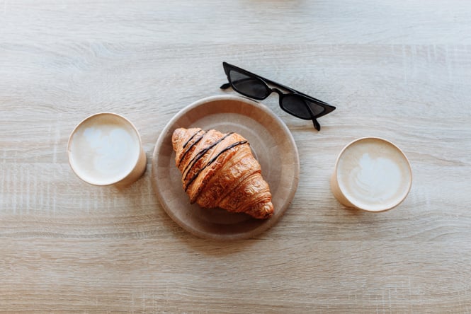 A croissant on a table sitting next to two coffees and sunglasses.