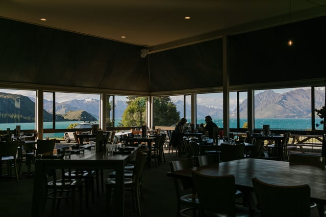 People dining inside a darkly lit restaurant with a lake and mountains in the background.