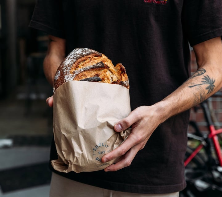 Man with tattooed arm holding a loaf of sourdough bread in brown paper bag