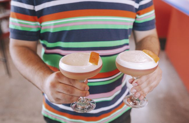 A wait staff in a bright stripe top holding two cocktails in his hands.