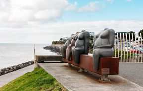 Four nude female sculptures sitting on a bench looking out towards the ocean.