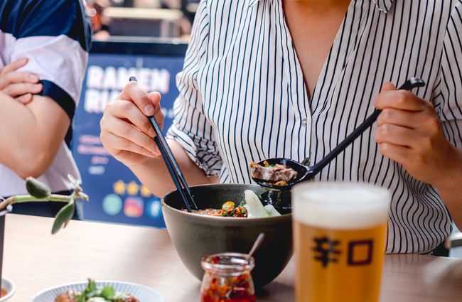 Woman eating a bowl of ramen with pint of beer.