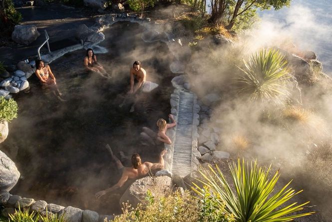 5 people sitting in a steaming hot pool surrounded by rocks and bushes.