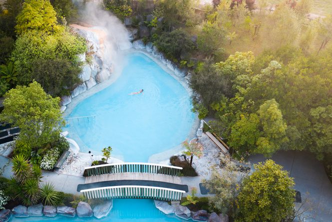 Bird’s eye view of a bright blue steaming hot springs with a bridge walkway going over the pool.