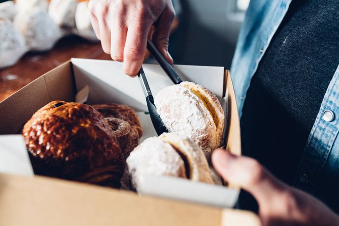 A person using tongs to dish donuts into a take away box.